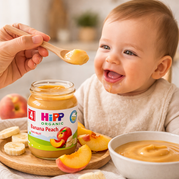 Baby being fed HiPP Organic Banana Peach baby food with a spoon, surrounded by fruit and a jar of the product.
