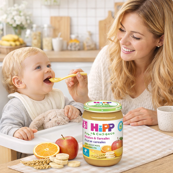 Woman feeding a baby with a spoon in a kitchen setting, with a Hipp baby food jar on the table.