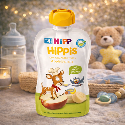 Hippis Apple Banana pouch with a deer illustration on a wooden surface with toys and a teddy bear in the background.