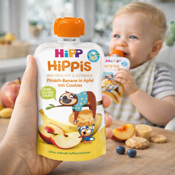 Hand holding a Hipp HiPPiS fruit and grain pouch with a baby in the background eating another pouch.