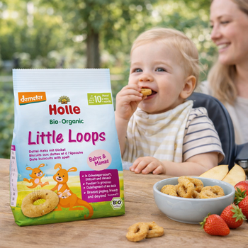 Baby eating Holle Organic Little Loops with a bowl of cookies and strawberries on a wooden table.