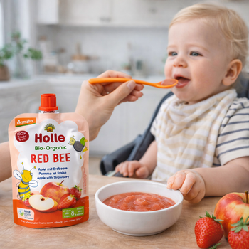 Baby being fed Holle organic red bee baby food with a spoon, surrounded by strawberries and an apple.