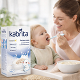 Woman feeding a baby with a bowl of cereal next to a box of Kabrita cereal.