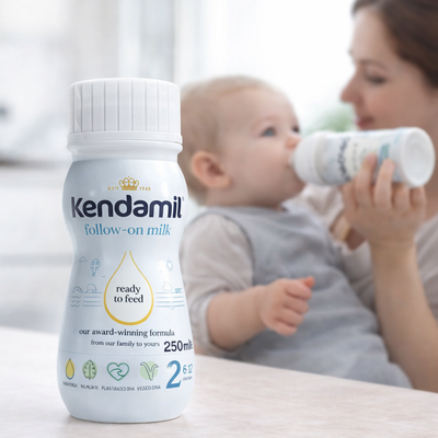 Kendamil follow-on milk bottle with a baby and mother in the background