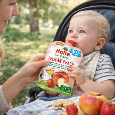 Baby in a stroller being fed Holle Pelican Peach organic baby food in a park setting.