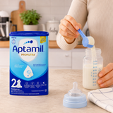 A person in a beige sweater is pouring milk from a blue scoop into a baby bottle on a kitchen counter, with a large Aptamil Pronutra powder canister standing beside the bottle.