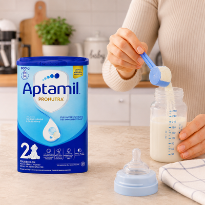 A person in a beige sweater is pouring milk from a blue scoop into a baby bottle on a kitchen counter, with a large Aptamil Pronutra powder canister standing beside the bottle.