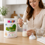 A woman in a cream cardigan is preparing baby formula, scooping milk from a bottle with a spoon next to a large canister of Hipp Anti-Reflux Infant Formula. The scene is set in a bright, modern kitchen with a wooden countertop.