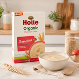 A box of Holle Organic Semolina Wholegrain Cereal by demeter, styled on a kitchen counter next to a bowl of creamy cereal, a wooden spoon, wheat stalks, and a jar of oats.
